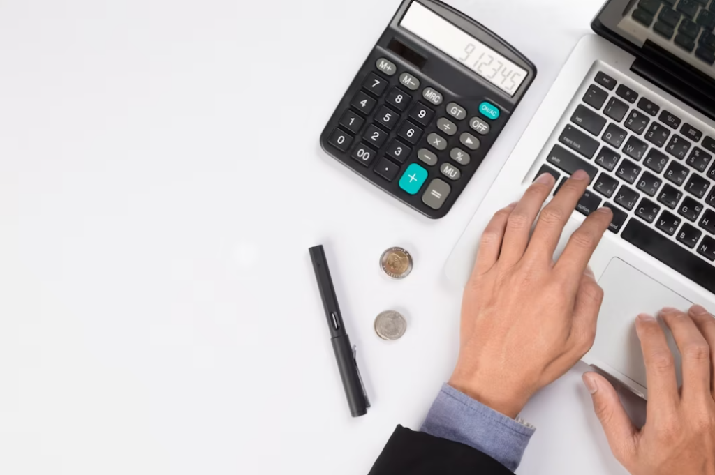 person is typing on a laptop, on the left on the table is a calculator, pen, and coins